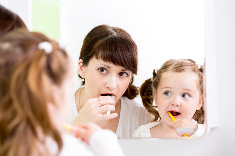 Mom and baby brushing teeth together in bathroom mirror.