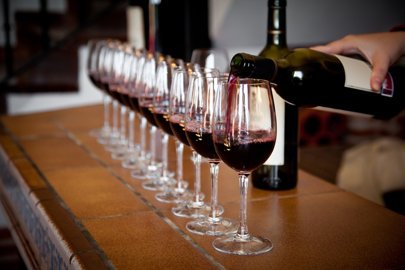 Pouring bottle of red wine into line of glasses on bar.