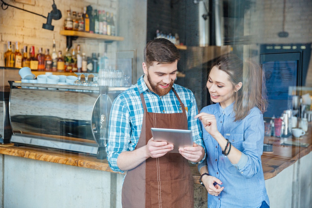 Smiling handsome waiter holding tablet and young pretty woman pointing on it in coffee shop.jpeg